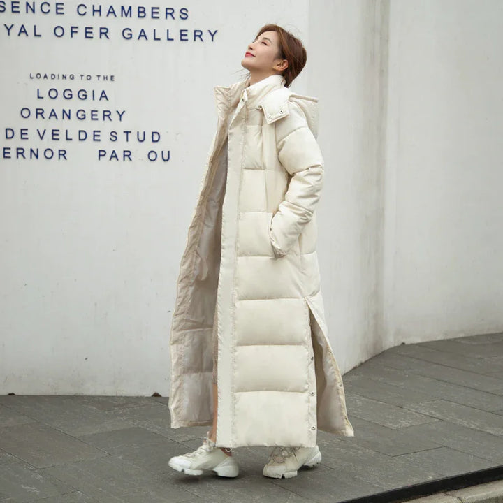 Woman wearing a long cream puffer coat and white sneakers standing against white wall with black text