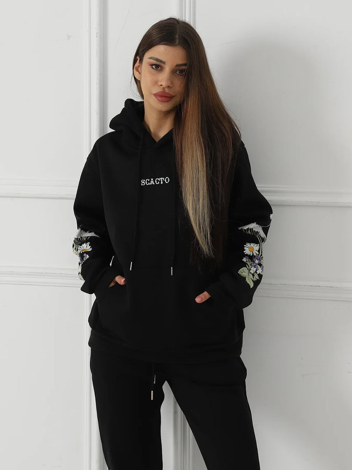 Woman wearing black hoodie with floral embroidery on sleeves and white text, standing against white wall