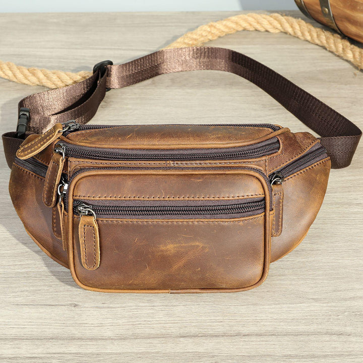 Brown leather waist bag with multiple zippered compartments and adjustable strap on wooden surface
