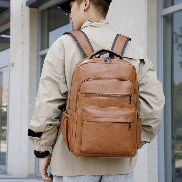 Person wearing tan leather backpack with multiple zippered pockets in urban setting