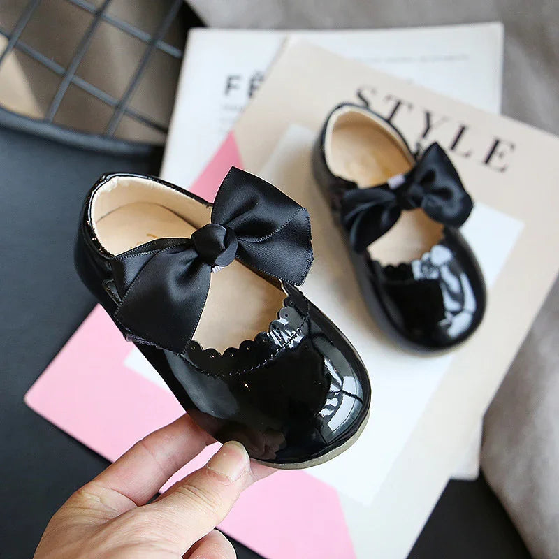 Black patent leather baby shoes with satin bow, held over fashion magazines