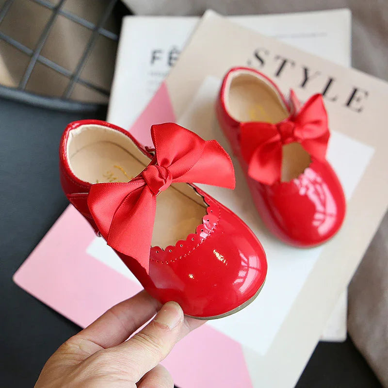 Hand holding shiny red baby shoes with large satin bows, placed on fashion magazines