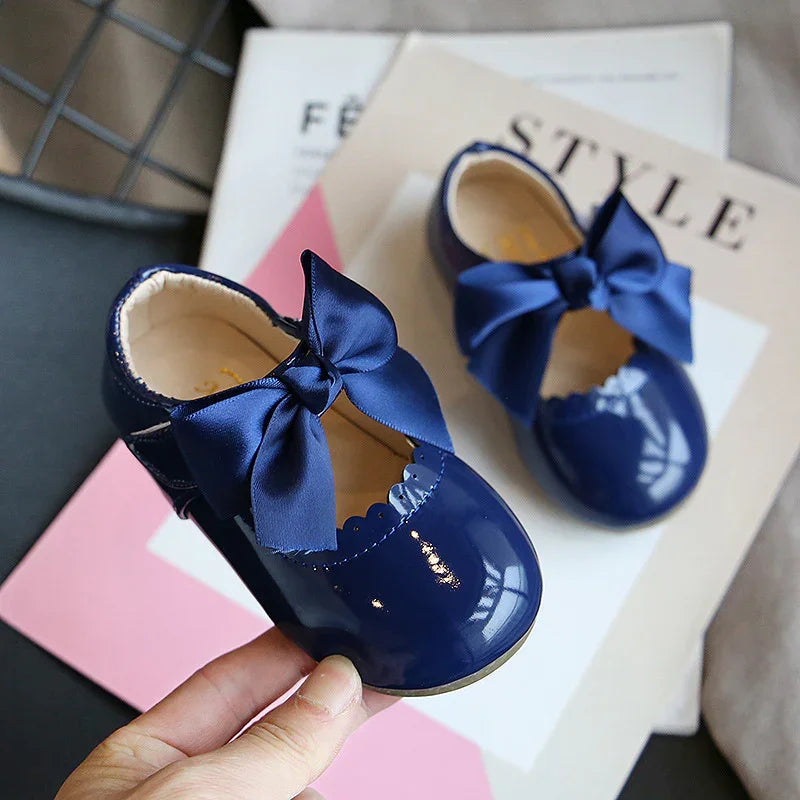 Pair of shiny navy blue baby shoes with large satin bows on top, held over fashion magazines