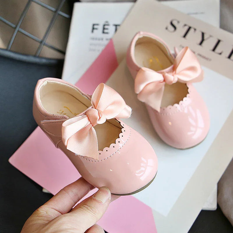 Pair of shiny pink baby shoes with large satin bows on top, displayed on styled magazines