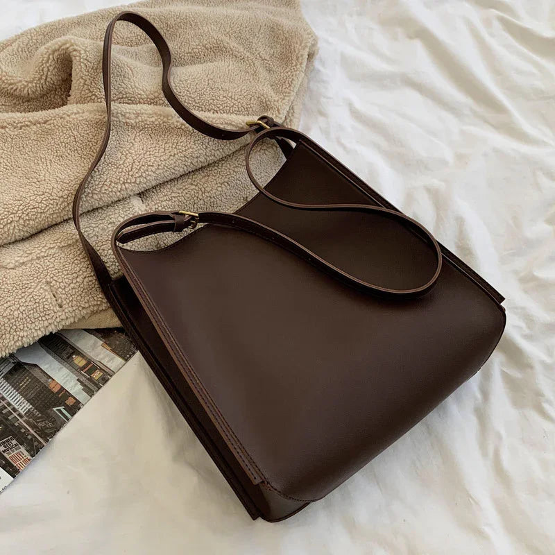 Dark brown leather handbag with long strap on white fabric next to beige sherpa jacket and magazine