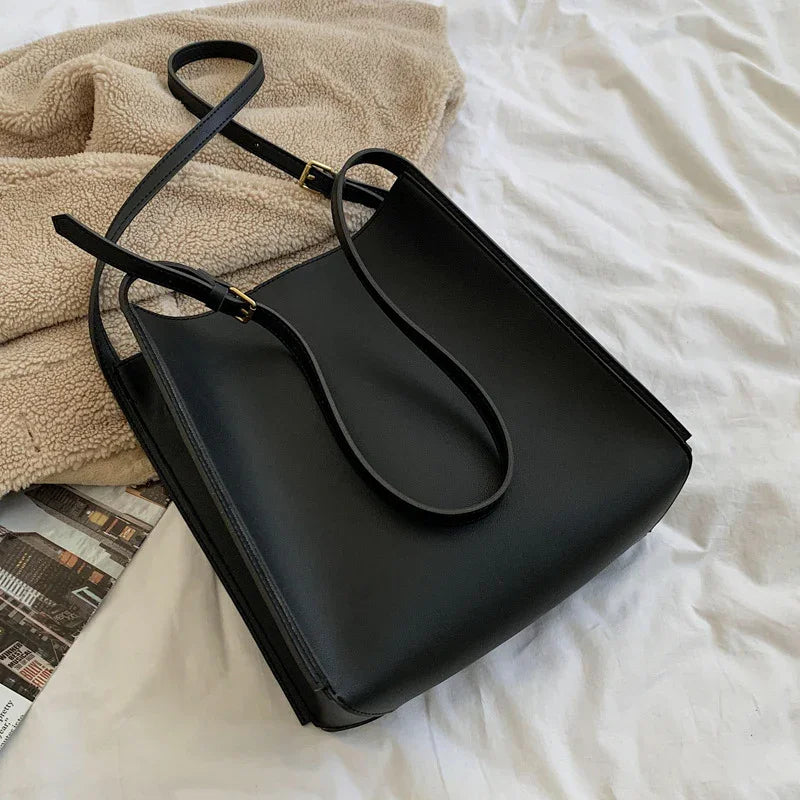 Black leather tote bag with adjustable straps on beige fleece blanket and white bed linens