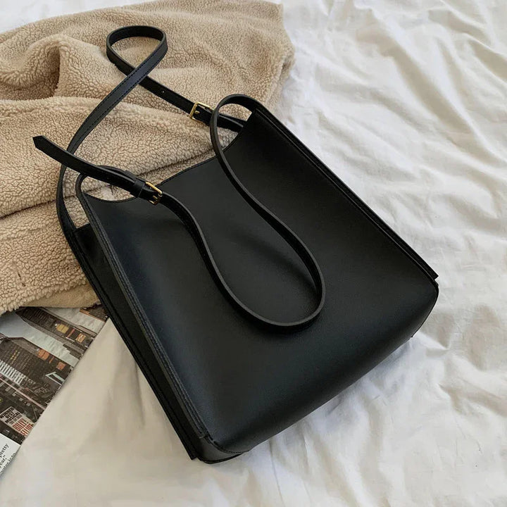 Black leather tote bag with adjustable straps on beige fleece blanket and white bed linens