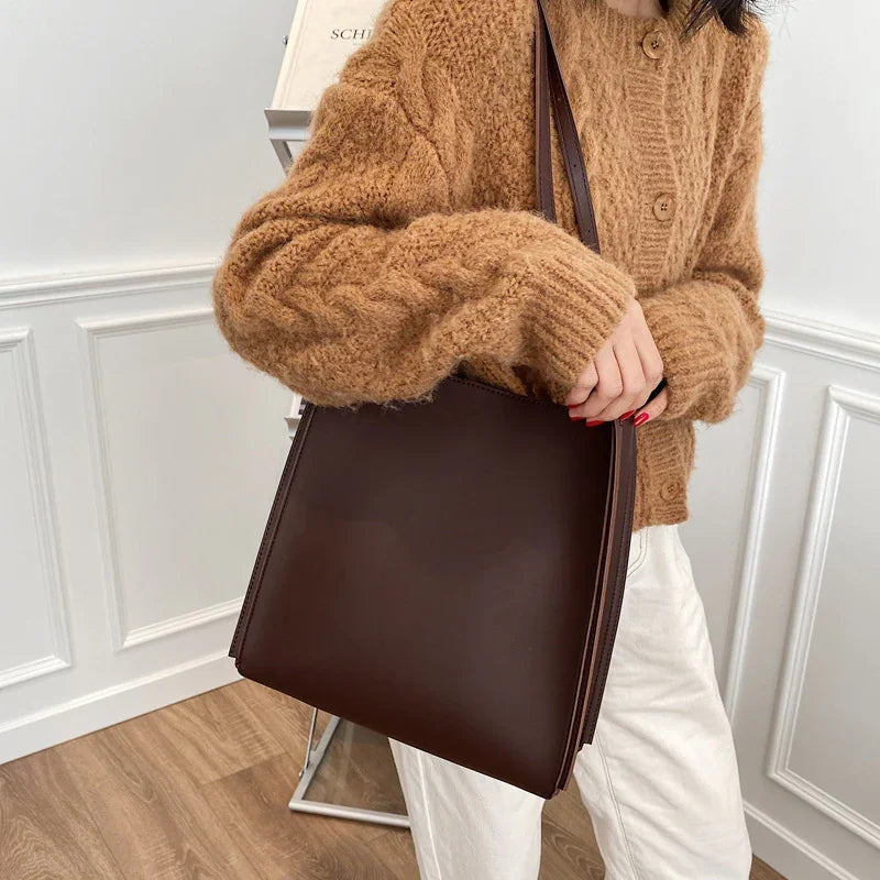 Woman wearing a brown fuzzy cardigan and white pants holding a dark brown leather tote bag indoors