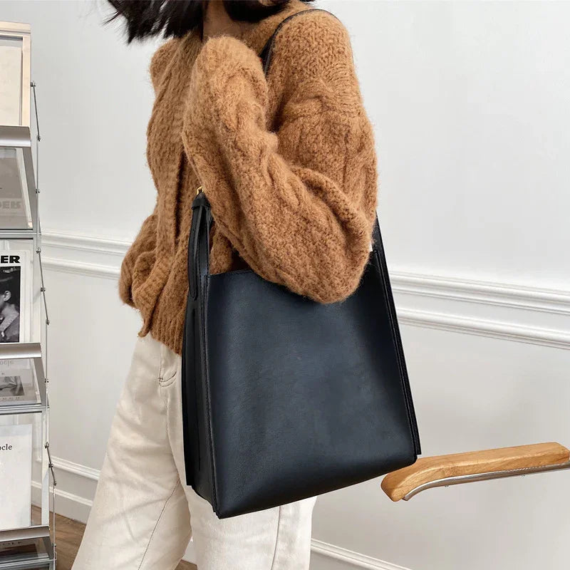 Woman wearing a brown fuzzy sweater and white pants carrying a black leather tote bag indoors