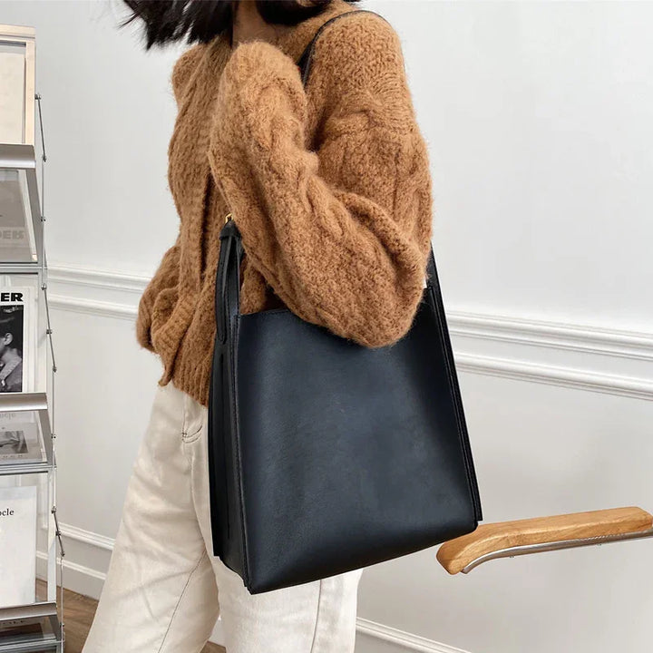 Woman wearing a brown fuzzy sweater and white pants carrying a black leather tote bag indoors