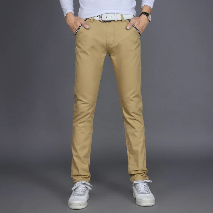 Man wearing beige slim-fit chinos with white belt and gray sneakers on gray background