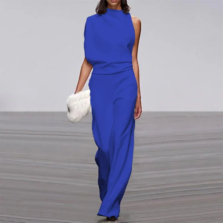 Model wearing royal blue sleeveless jumpsuit with one-shoulder design walking on runway holding white clutch