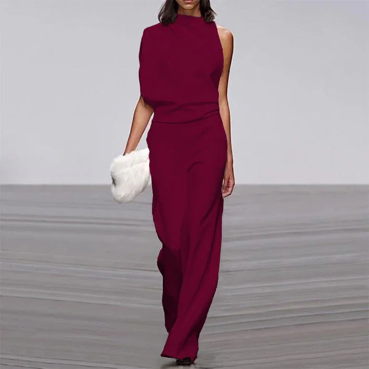 Model walking runway in sleeveless asymmetric burgundy jumpsuit holding white fur clutch