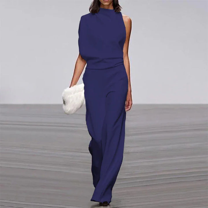 Model wearing a navy blue sleeveless asymmetrical jumpsuit holding a white fur clutch on runway