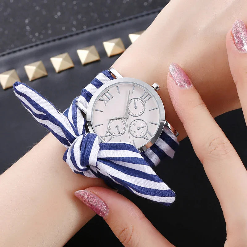 Silver wristwatch with white dial and Roman numerals on wrist, featuring a blue and white striped fabric strap tied in a bow
