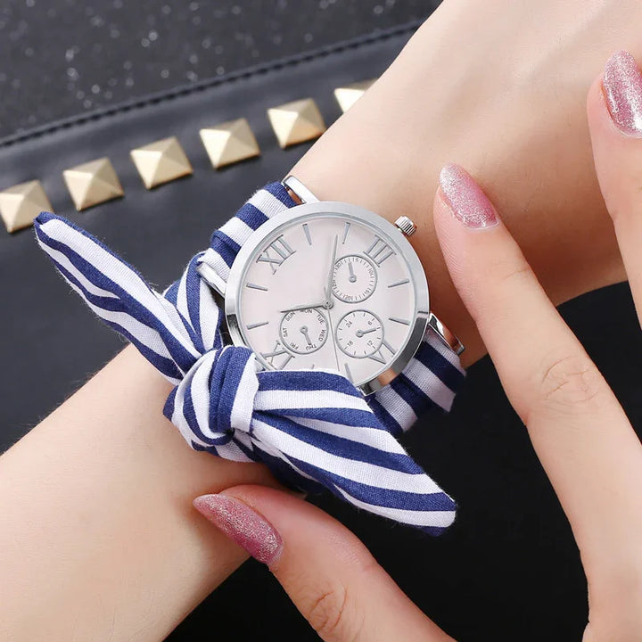 Silver wristwatch with white dial and Roman numerals on wrist, featuring a blue and white striped fabric strap tied in a bow
