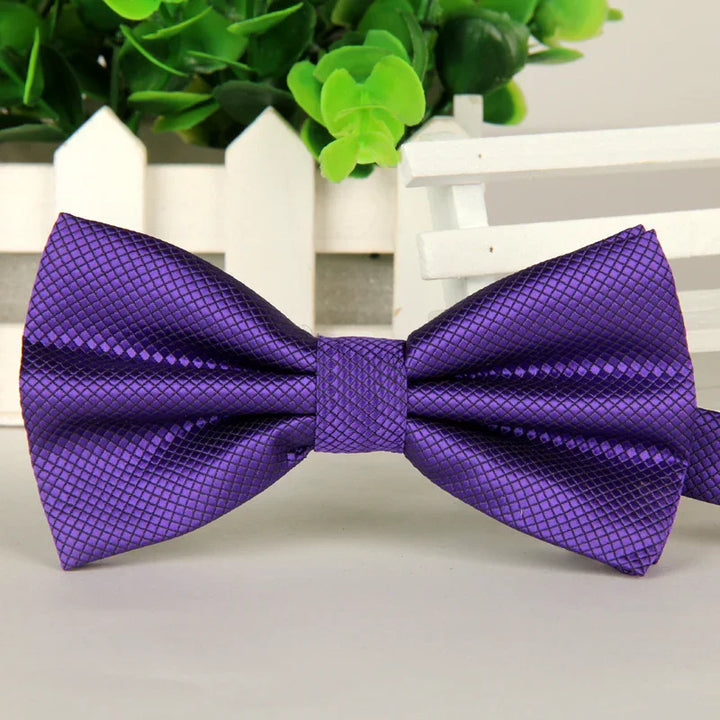 Purple textured satin bow tie displayed on white surface with green leaves and white picket fence background