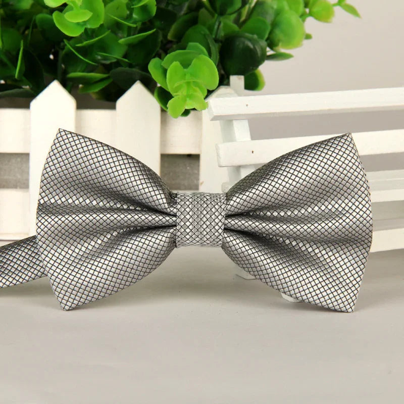Silver textured men's bow tie with black grid pattern on white surface with green plant background
