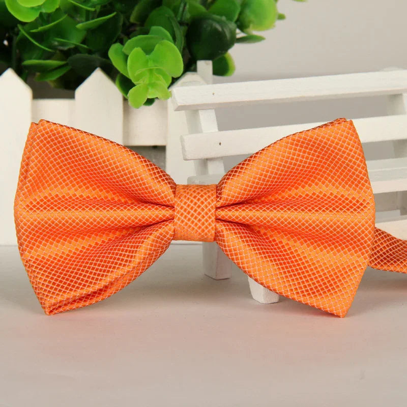 Bright orange silk bow tie with a subtle grid pattern on a white surface with green plant and white wooden fence background