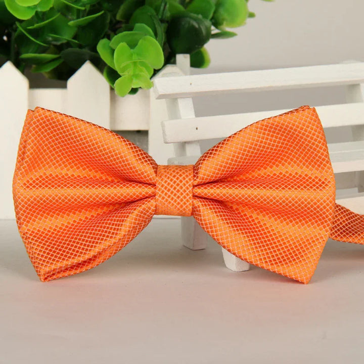 Bright orange silk bow tie with a subtle grid pattern on a white surface with green plant and white wooden fence background