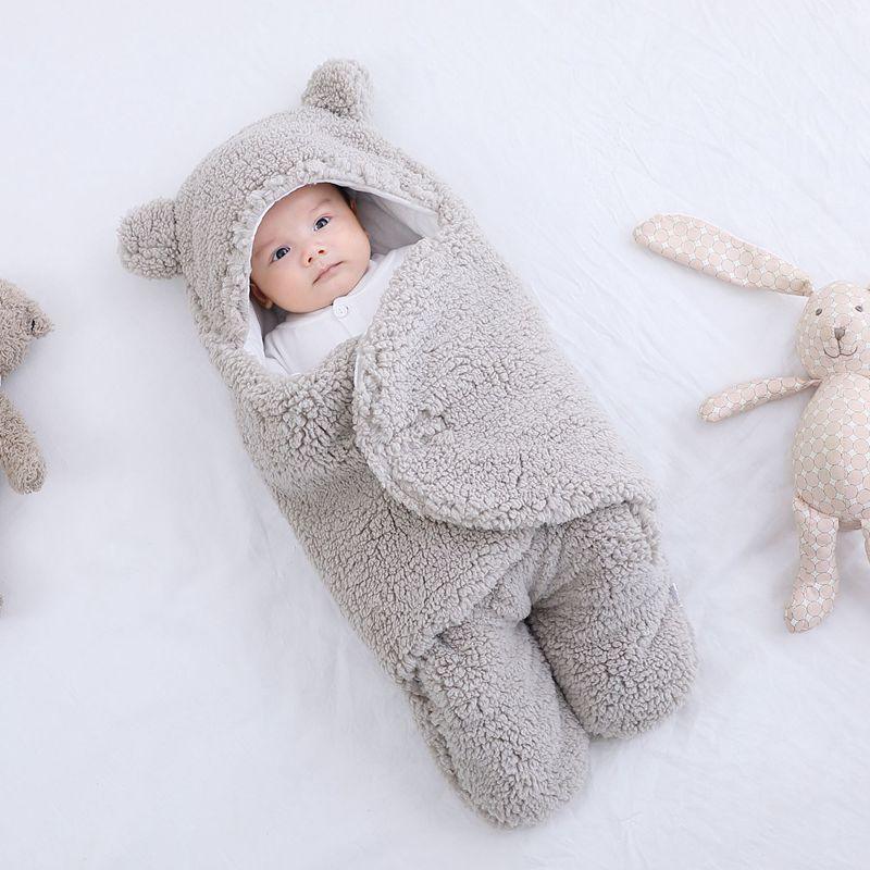 Baby wrapped in soft gray bear-themed fleece swaddle blanket lying on white bed near stuffed animals