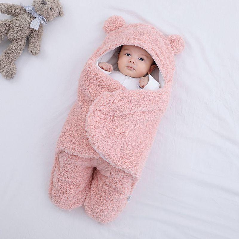 Baby wrapped in a soft pink bear-themed fleece swaddle blanket lying on a white surface with a teddy bear nearby