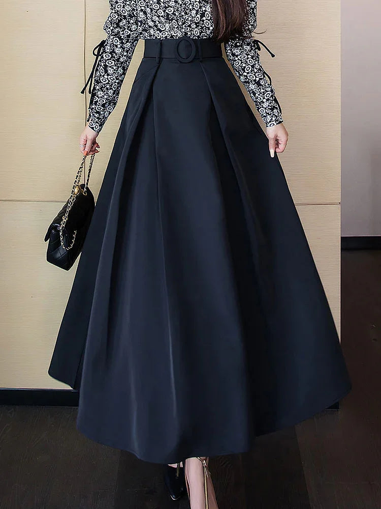 Woman wearing a high-waisted black A-line maxi skirt with belt, paired with a floral long-sleeve blouse and holding a black handbag