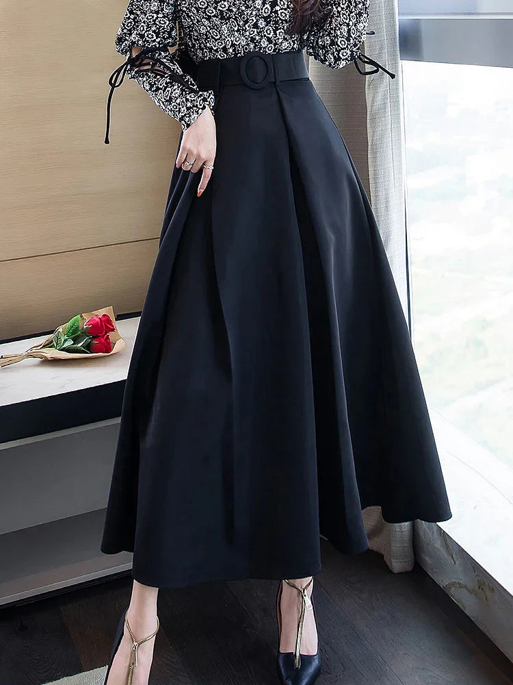 Woman wearing high-waisted black midi skirt with buckle belt and black floral blouse by window