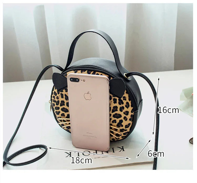 Small round leopard print crossbody bag with black ears, whiskers, and adjustable strap on white table