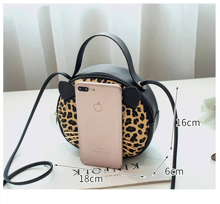 Small round leopard print crossbody bag with black ears, whiskers, and adjustable strap on white table