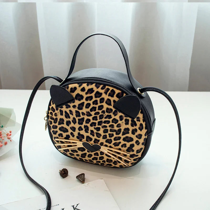 Round leopard print crossbody bag with cat ears, whiskers, and heart-shaped nose on white table