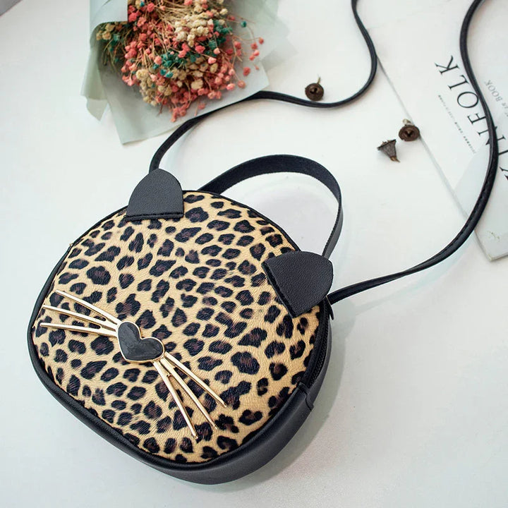 Leopard print cat-shaped crossbody bag with black ears and whiskers on white surface with dried flowers