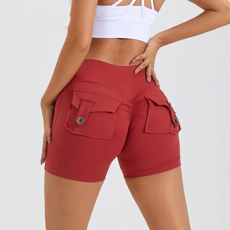 woman model wearing rust red high-waist ruched booty shorts with flap button pockets and white sports bra
