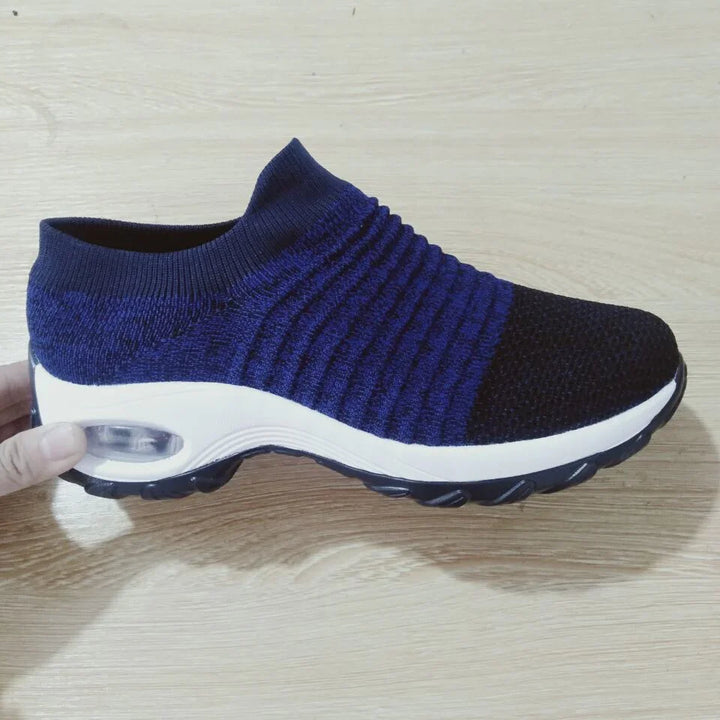 Hand holding a navy blue and black knitted slip-on sneaker with white thick sole on wooden floor