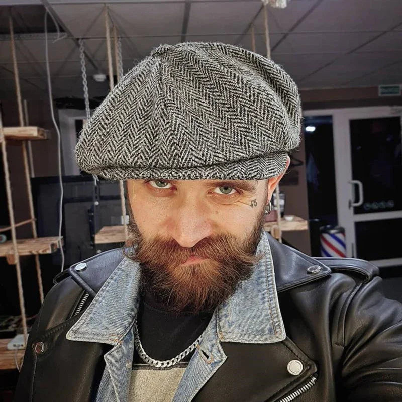 Man with beard wearing a herringbone newsboy cap, leather jacket, and denim collar in indoor setting