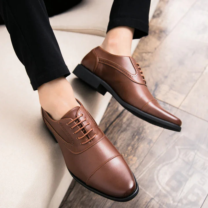 Brown leather men's dress shoes with cap toe and laces on wooden floor and beige chair
