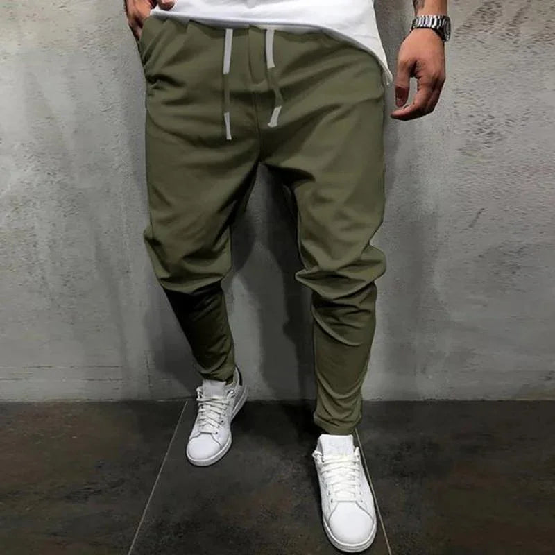 Man wearing olive green jogger pants with white drawstrings and white sneakers against gray wall