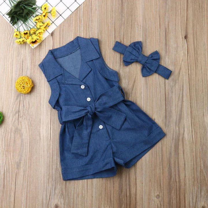 Blue sleeveless toddler romper with buttons and bow belt, paired with matching blue headband on wooden surface