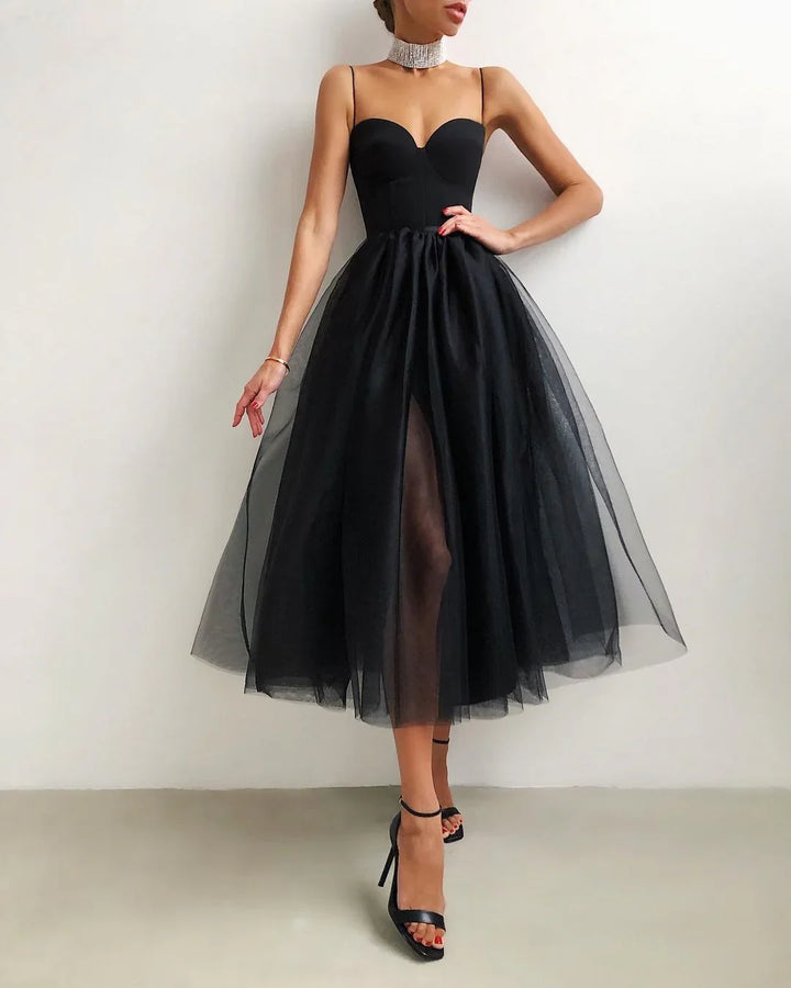 Woman in elegant black tulle midi dress with slit, strappy heels, and rhinestone choker against white wall