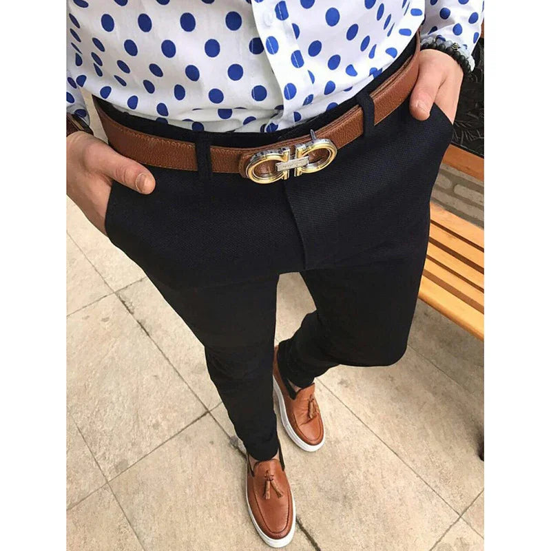 Man wearing black pants, brown leather belt with gold buckle, white shirt with blue polka dots, and brown tassel loafers