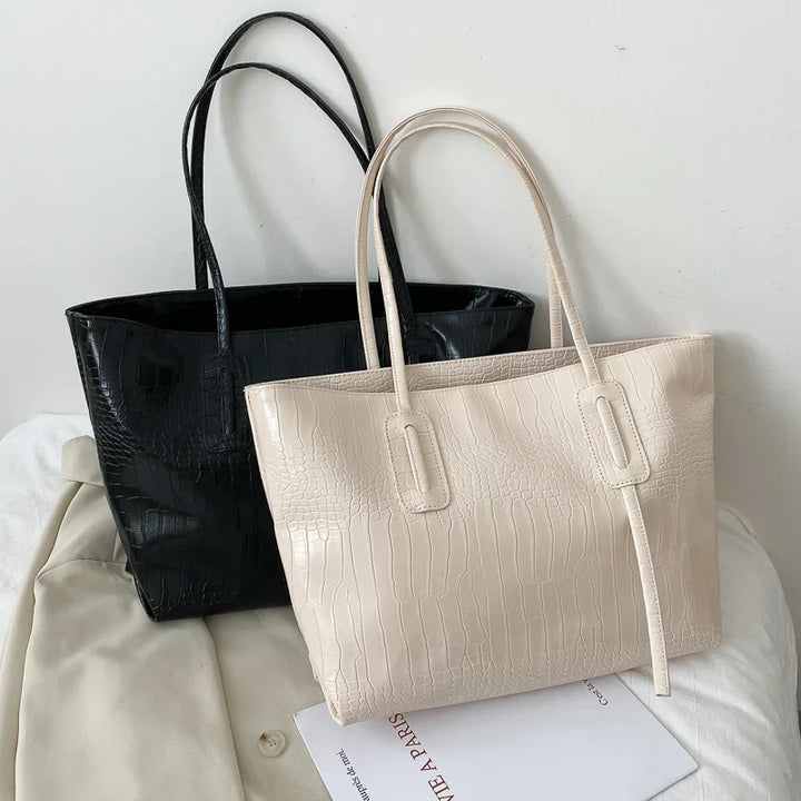 Two crocodile embossed leather tote bags, one black and one beige, displayed on white fabric and magazine
