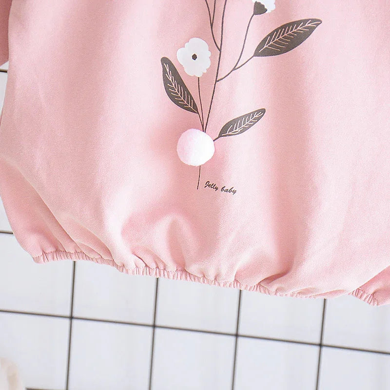 Pink baby romper with flower print, pom-pom detail, and elastic hem on tiled background