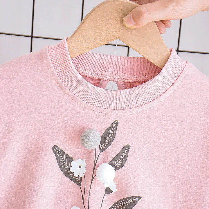 Close-up of pink sweatshirt with floral design and pom-poms on wooden hanger