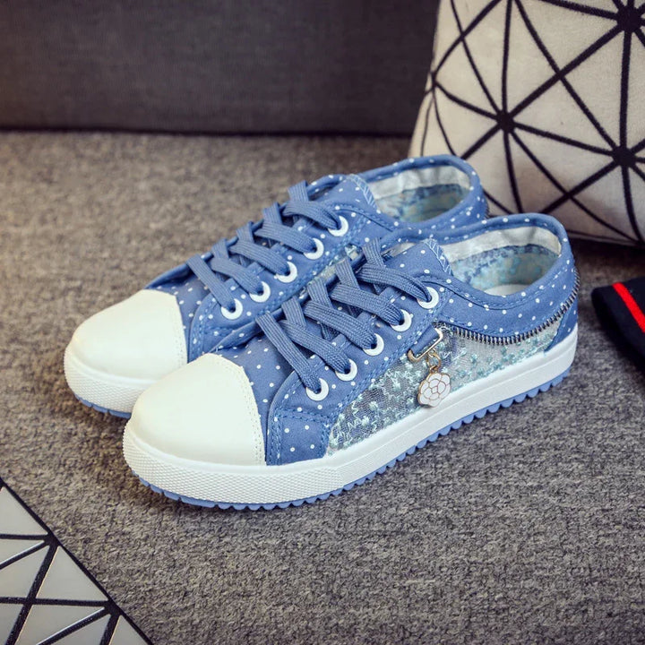 Blue polka dot canvas sneakers with white rubber toe caps and decorative flower charm on gray carpet
