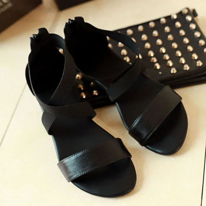 Black leather flat sandals with ankle straps and open toes on beige surface next to studded black clutch