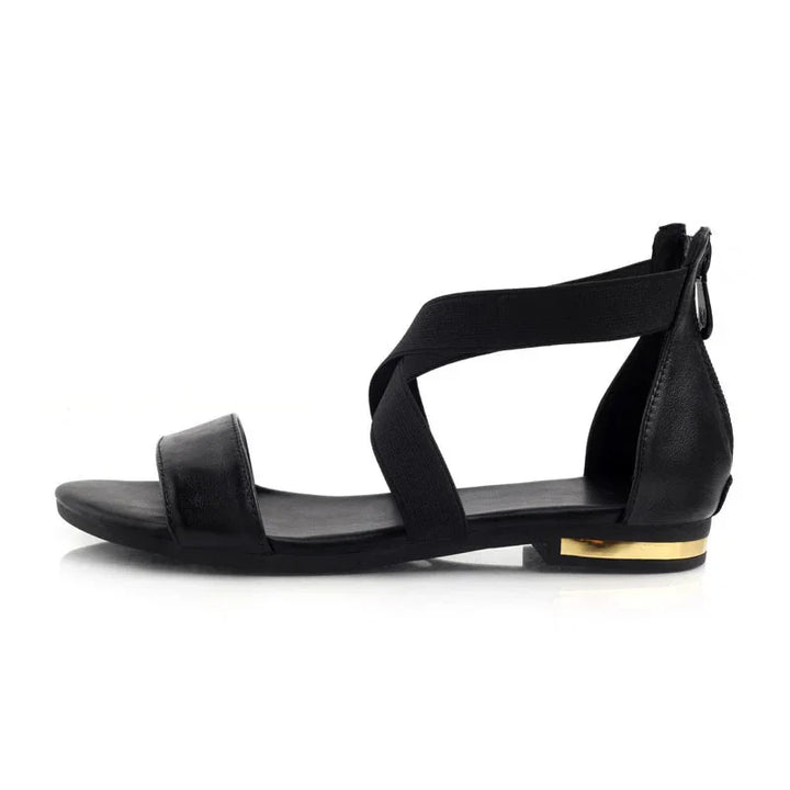 Black flat sandal with elastic cross straps and gold accent on the heel, isolated on white background