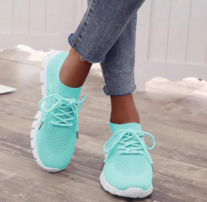 Close-up of feet wearing turquoise knit sneakers with white soles and light blue jeans on wooden floor
