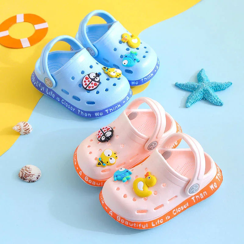Blue and pink kids' clogs with colorful animal charms and text on soles, beach-themed background