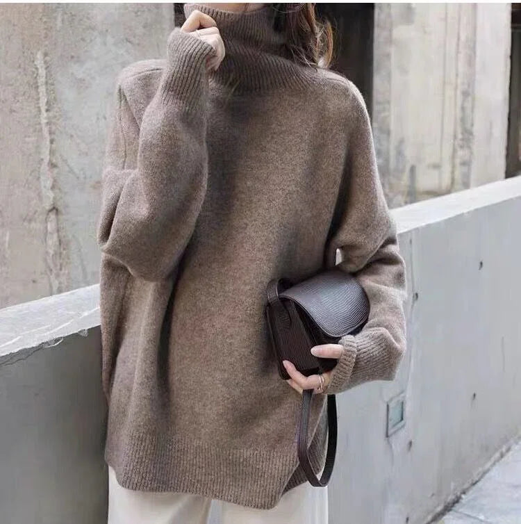 Woman wearing oversized brown turtleneck sweater holding small dark leather crossbody bag outdoors