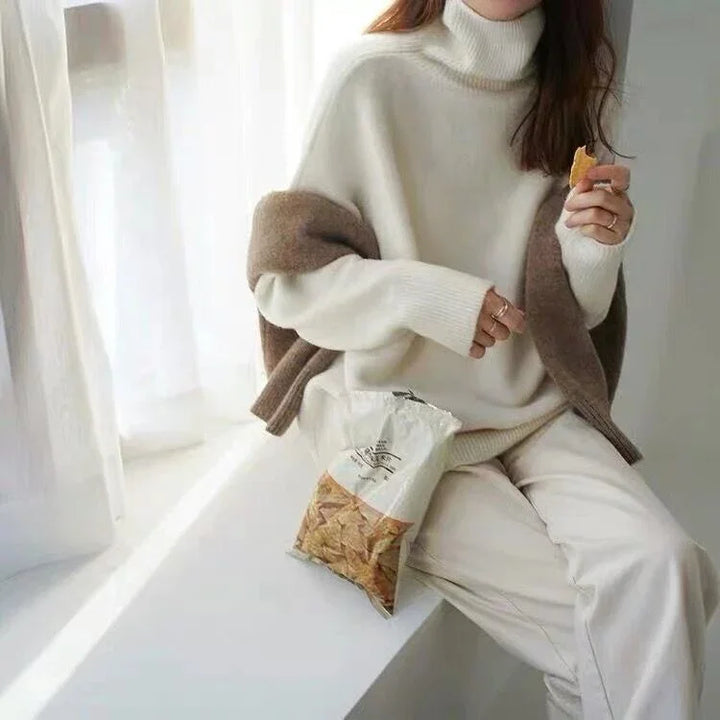 Woman wearing cream turtleneck sweater and beige pants sitting by window eating chips from a bag
